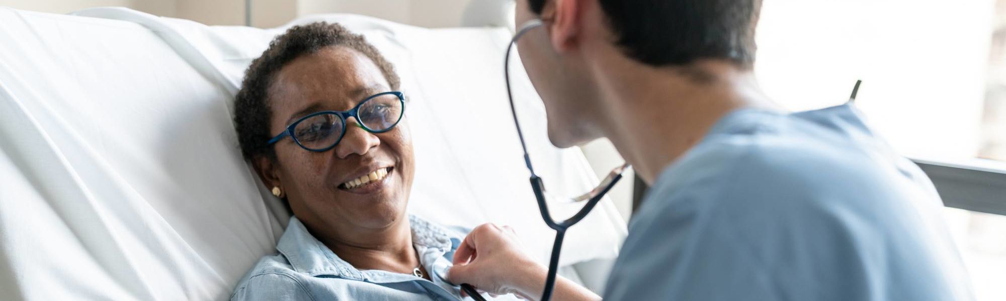 Female black patient lying down on hospital bed while nurse is checking her heart beat with stethoscope